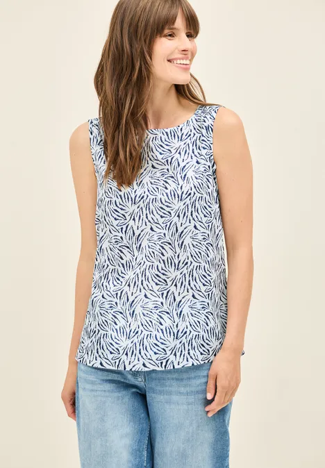 Frau mit gemustertem ärmellosen Top und Jeans lächelt.